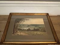 Framed watercolour painting featuring a pastoral landscape scene with sheep, trees and hills under a cloudy sky. The painting is matted with a beige mat and enclosed in an ornate gold frame.