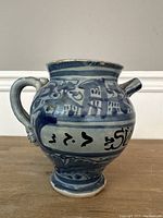 Front side of antique blue and white ceramic vessel with handle, showing blue painted floral patterns and a section of black text on white band.