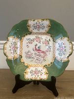 Full view of antique Minton's plate standing on display with green background, floral motifs, and phoenix central design.