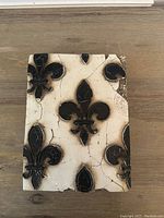 Front view of the rectangular ceramic tile featuring several black Fleur de Lis motifs on a cream background with visible cracks and aging.