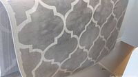 Rolled and unrolled views of a large grey and white patterned area rug with geometric trellis design.