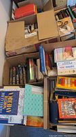 Photo showing multiple cardboard boxes filled with various hardcover and paperback books including fiction and travel guides.