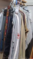 Side view of men's clothing hanging in closet including a grey athletic jacket, beige trench coat, and striped shirts. Also visible are several ties including one woven colorful tie.