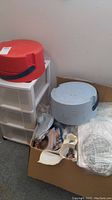 White plastic storage drawers with red rotating marker holder on top and blue rotating holder next to it. Cardboard box on floor containing craft supplies and plastic items.