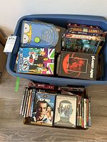 Top-down view of a large bin containing various DVD movies and TV shows, with some DVDs laid out underneath the bin. Visible titles include The Simpsons Season 7, Planet of the Apes, Smallville Season 5.