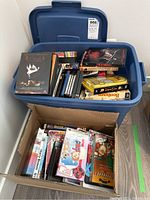 Blue plastic bin filled with assorted DVDs and VHS tapes, with a smaller box in front containing additional movie discs and cases.