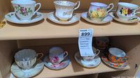 Eight fine china teacups and matching saucers arranged on wooden shelving, some with floral and gold designs.