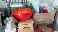 View of plastic mixing bowls nested inside each other in red, orange, yellow, and green colors with plastic storage containers and kitchen utensils in a cardboard box.