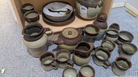 Wide view of entire lot showing collection of pottery pieces arranged on floor and shelf corner. Includes jugs, mugs, bowls, goblets and lidded pot in natural earth glazes.