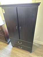 Full front view of the wood storage cupboard showing two upper doors with keyhole handles and two drawers below with metal handles. Dark brown color and smooth finish.