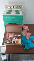 Photo showing teal Lady Homestead toy oven with four burners and oven door. Pink and blue plastic toy dishes and cups arranged next to oven. Box containing metal baking molds and toy utensils in front.