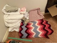 White bedding sheets and pillowcases, red checkered fabric, crocheted afghan, and pink striped sheets laid in a pile with crocheted afghan underneath.