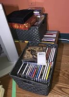 Black woven media storage baskets containing multiple CDs and cassettes on wooden floor against peach wall.