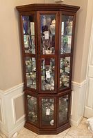 View of the wood finished corner china cabinet with glass doors and shelves displaying various items with internal light on.