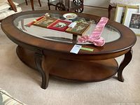 Front angle of round coffee table showing wood frame, glass oval top, curved legs, and bottom shelf. Items on table are visible but not included.