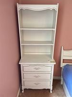 Front and side view of white three-drawer dresser with ornate metal pulls and removable two-shelf hutch with scalloped trim.
