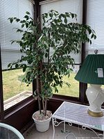 Full view of the faux ficus tree in a white plastic pot placed in corner of porch with blinds in background.