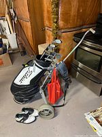 Full view of golf bag on push cart with golf clubs and golf shoes placed nearby on floor.