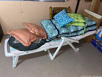 Resin lounge chair with various cushions stacked on top including orange cushions, blue paisley cushions, white and blue geometric pillow, and green cushion