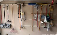 Wide shot of neatly hung yard tools on wall rack in garage including shovel, rakes, brooms, extension cord, and more.