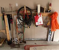 Garage wall with various garden and yard tools hanging including rake, pick axe, axe, extension cord, garden gnome, orange raincoat, and other hand tools.