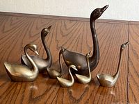 Seven brass swan figurines in varying sizes and poses lined up on wooden surface; tallest measures 8 inches. Shows overall lot contents and scale.