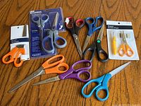 Ten pairs of scissors on wooden surface including some in original packaging and various handle colors and sizes.