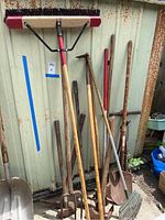 Wide view of all yard maintenance tools leaning against a metal shed showing handles and tool heads