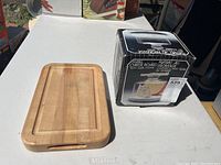 Wooden cutting board with slot handles, lightly used, placed next to box of marble cheese board with glass dome.