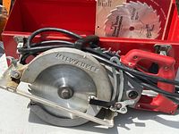 Milwaukee worm drive circular saw with blade attached and power cord, resting in red metal storage box. The saw has a metal blade guard and red ergonomic handle.