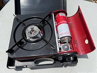 Photo showing portable single burner gas stove with black grate and exposed gas canister compartment in red, inside plastic carrying case.