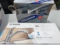 Two boxed Moen kitchen faucets, one still shrink-wrapped and the other in original packaging with product images and features visible on the box.