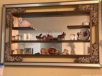 Full view of large vintage gold-tone framed mirrored wall shelf with various decorative porcelain and ceramic figurines arranged on three metal shelves