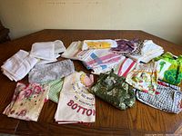 Overview of multiple white towels, patterned cloths, aprons, and fabric pieces laid on a table.