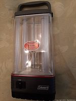 Front view of Coleman battery lantern showing transparent casing, black base with red accent, handle, on/off switch, and Coleman logo.