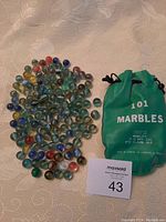 Image showing a collection of assorted glass marbles and a green drawstring bag labeled '101 Marbles'. Marbles include various colors such as blue, red, yellow, and clear with swirl patterns.