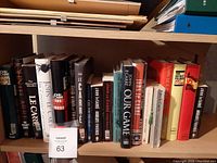 View of multiple John LeCarre and related books on a shelf, showing a mixture of hard and soft cover books mainly in darker color tones