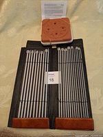 Set of 12 metal knitting needles organized in two rows, size markers visible. Laid on a black leather case backing surface, with a flower press and information paper behind.