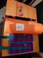 Overview photo showing orange yoga mat, black and red muscle roller stick, pink 2lb hand weight dumbbell, and Weider leg weights on wooden surface.