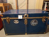 Front and top view of blue metal trunk with brass edges and two latch locks, central lock, stickers on front including a deer and moon sticker, and signs of wear.
