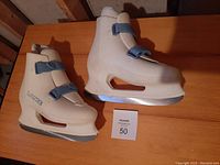Pair of white molded plastic Lange figure skates with blue buckles and silver blades shown from front angle on a wooden surface under indoor lighting.