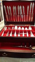 Full set of silverplate cutlery displayed in open wooden case lined with red fabric, showing knives, spoons, and forks arranged in compartments.