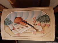 Close-up of top of cream wooden trunk showing handpainted violin, bows, sheet music, roses, and leaves.