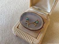 Front angled view of the oval chalcedony brooch showing silver gilt setting and applied foliate decoration with turquoise stones inside a vintage box.