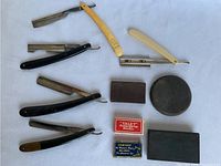 Five vintage straight razors positioned around three whet stones and two razor blade packages on a white surface.