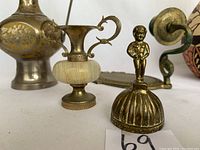 Close view of three brass items: a lidded urn-shaped container with two handles, a small bell with a figurine handle, and a brass item with a scrolled handle.