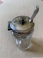 Top angled view showing the hinged silver-plated lid with finial and small silver-plated spoon inside the crystal condiment dish.