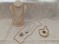 Full lot view showing three white and beige bead necklaces on stand and laid down, a rhinestone brooch, pearl necklace, and brooch with floral design.