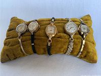 Front view showing all five vintage ladies watches on mustard colored velvet cushion, showing various gold and silver tone cases and straps, different dial shapes.