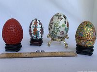 Four decorative eggs lined up side by side with a ruler showing sizes against a plain white background.